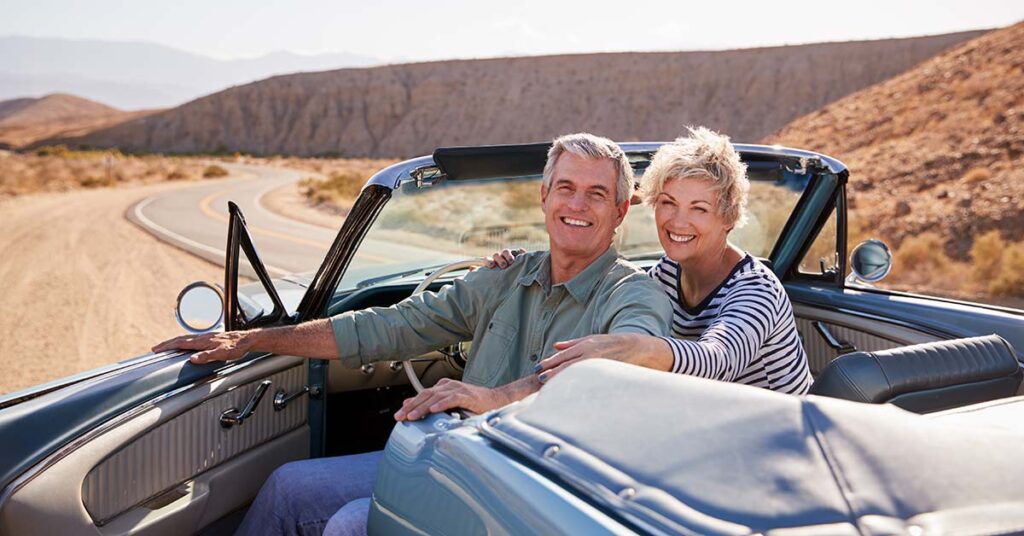 Ein Seniorenpaar sitzt in einem Cabrio auf einer Landstraße in einer weiten Landschaft, die Fahrertür ist auf und beide blicken nach hinten | Immobilienverrentung