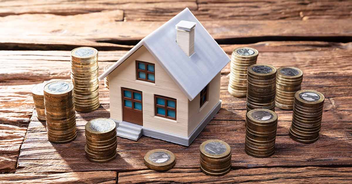 Modell eines Hauses steht auf einem Holztisch und darum stehen kleine Türmchen aus Ein-Euro-Münzen | Immobilienfinanzierung